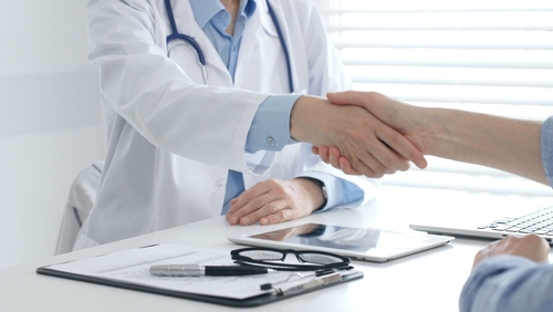 Doctor shaking patient’s hand across desk with tablet, clipboard, and stethoscope visible