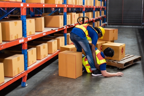 Philadelphia Warehouse Worker Injury Total Disability Workers Comp Warehouse worker clutching shoulder while coworker assists beside stacked boxes and pallet jack