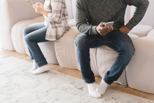 Man and woman sitting apart, each looking at smartphone on bed