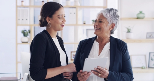 Two businesswomen in blazers smiling while reviewing tablet in office meeting