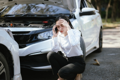 Woman crouching beside damaged car, speaking on phone with hood raised