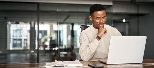 Professional man wearing glasses working at laptop beside open notebook in office