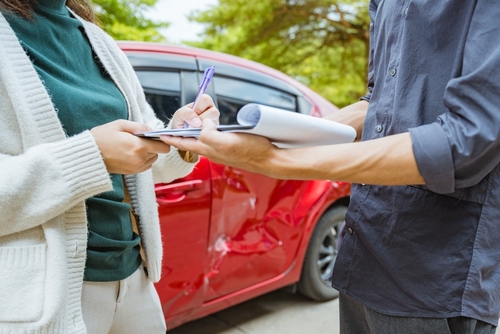 Woman signing clipboard beside dented car door after traffic collision