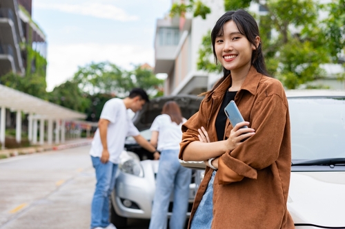 Happy Women Drivers After Brooklyn Car Accident No-Fault Claim Woman holding smartphone beside car while couple inspects open hood nearby