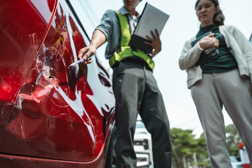 Insurance adjuster with clipboard pointing at dented car door beside driver
