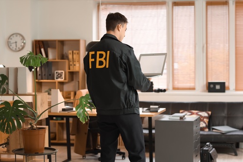 FBI jacket lettering visible as agent stands holding laptop in office