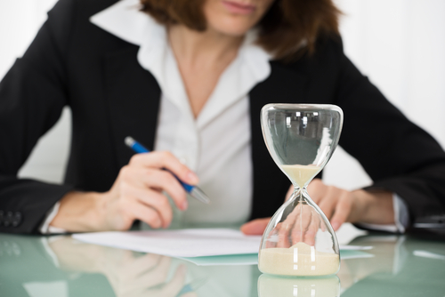 Hourglass in foreground beside businesswoman writing on document at desk