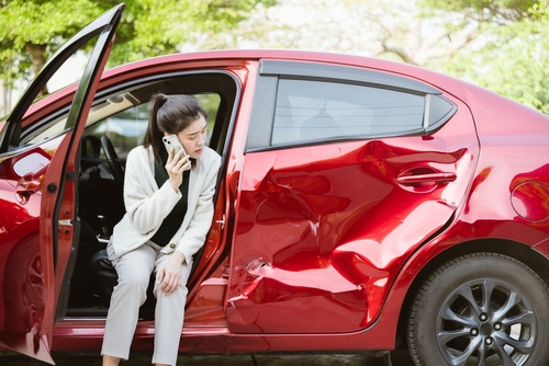 woman on phone sitting in open door of crash-damaged car