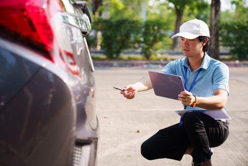 insurance adjuster crouching beside damaged vehicle holding tablet and pen