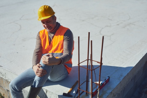 construction worker in hard hat and safety vest clutching injured knee on job site