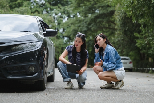 two women crouching beside damaged vehicle inspecting front end after collision