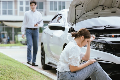distressed woman holding phone seated beside car with open hood after collision