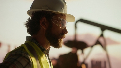 bearded oil worker wearing hardhat safety glasses and high-visibility vest at worksite