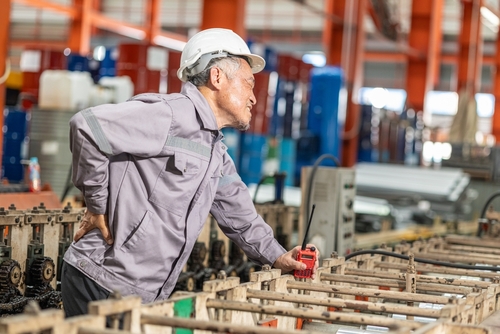 senior factory worker in hard hat clutching lower back in pain on industrial floor
