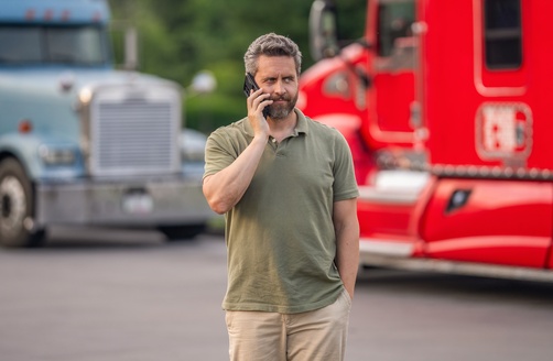 man talking on phone standing in front of semi trucks on road