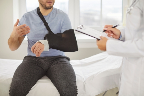 male patient wearing arm sling speaking with doctor holding clipboard