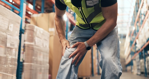 Las Vegas Warehouse Worker Knee Injury Workers Comp Claim warehouse worker in safety vest clutching knee in pain among stacked boxes