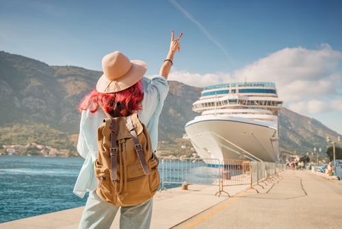 tourist wearing backpack and hat gesturing toward large cruise ship at port