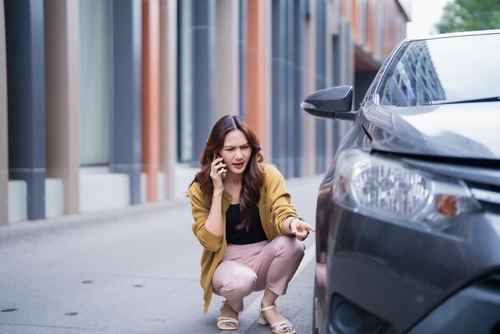 woman crouching beside damaged car bumper while talking on phone