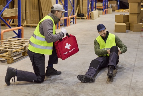 warehouse worker holding First Aid Kit assisting injured coworker on floor