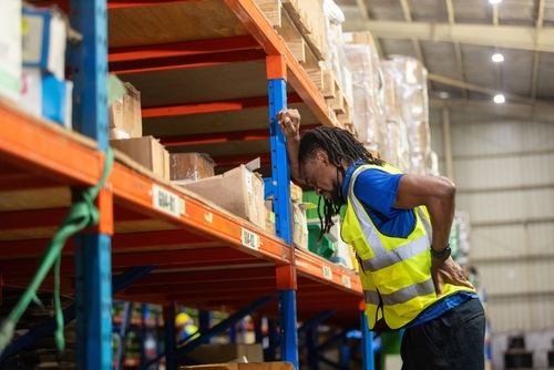 warehouse worker in safety vest clutching back near storage shelving