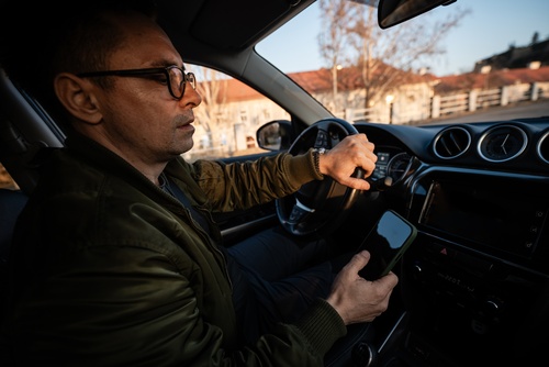 adult man holding smartphone while driving car with one hand on steering wheel