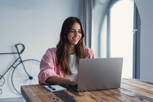 smiling young woman working on laptop at wooden desk near window