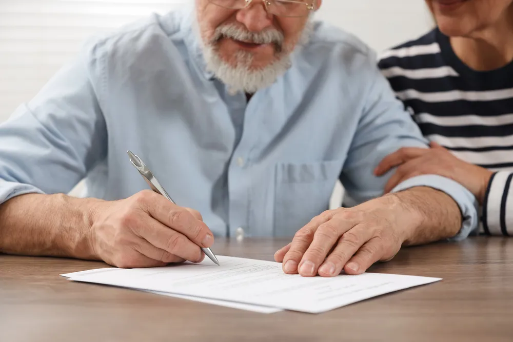 Older man signing document with pen as woman places hand on shoulder