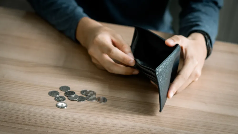 person holding open empty wallet beside scattered coins on wooden table